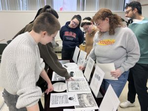 A group of Brock students stand with Professor of English Leah Knight around a table draped in a black table cloth displaying black and white text and visual images of historical materials. The students and professor smile as they look at the materials together.