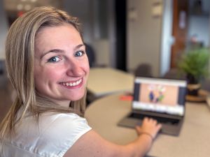 A young woman looks over her shoulder with a smile while sitting at a table using a laptop.