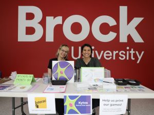 Two women sit behind a table with gaming information on it, in front of red wall with white lettering that says, Brock University.