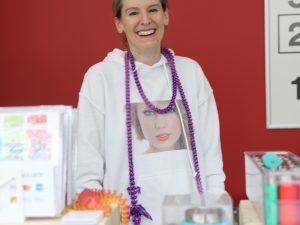 A women wearing a Taylor Swift sweater and a purple beaded necklace smiles while standing behind a table of items.