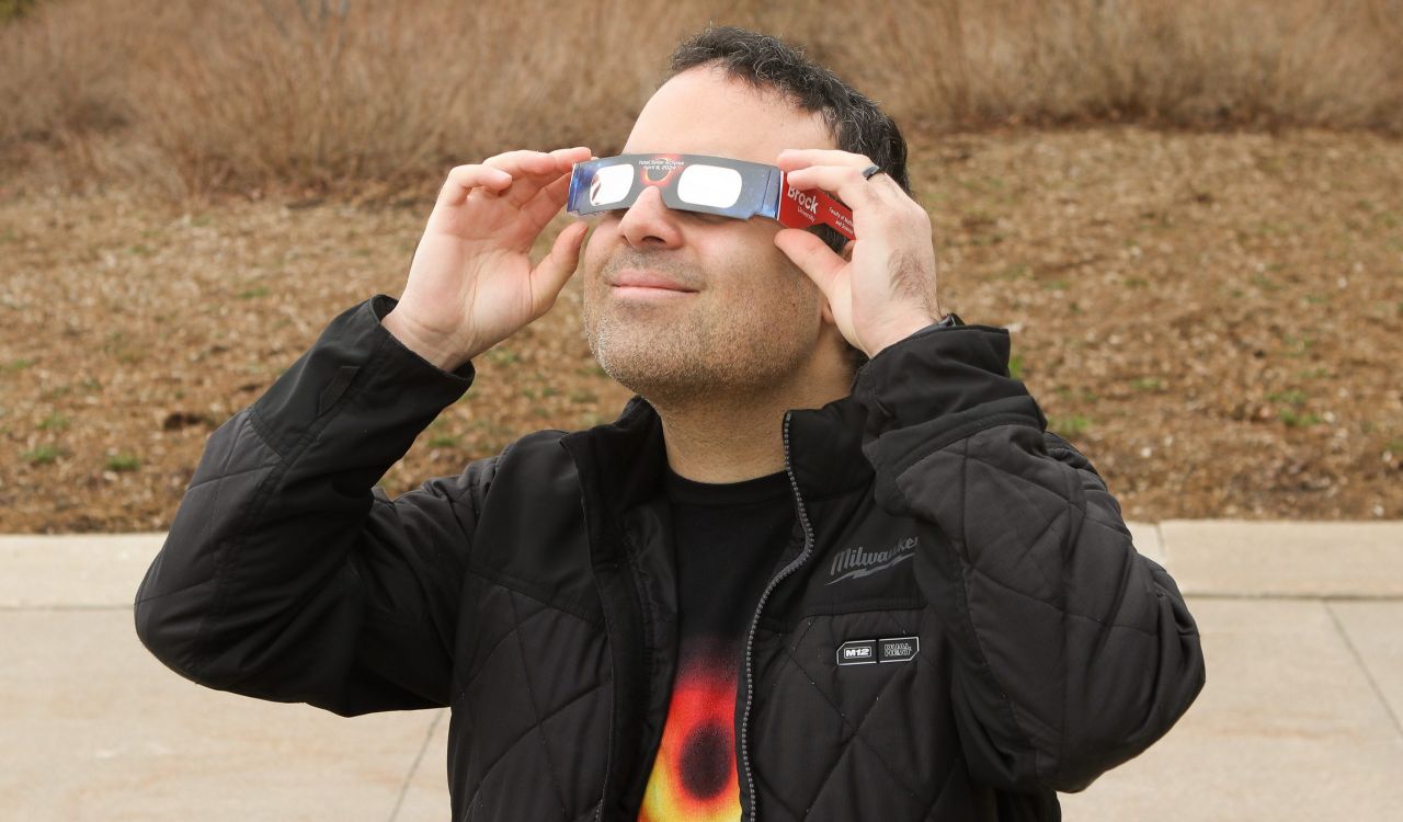 Physics professor Barak Shoshany holds a pair of Brock University branded solar eclipse viewing glasses to his eyes. He is wearing a T-shirt with an illustration of a wormhole, the hypothetical passage through space-time.