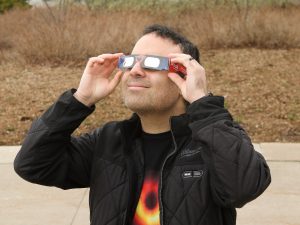 Physics professor Barak Shoshany holds a pair of Brock University branded solar eclipse viewing glasses to his eyes. He is wearing a T-shirt with an illustration of a wormhole, the hypothetical passage through space-time.