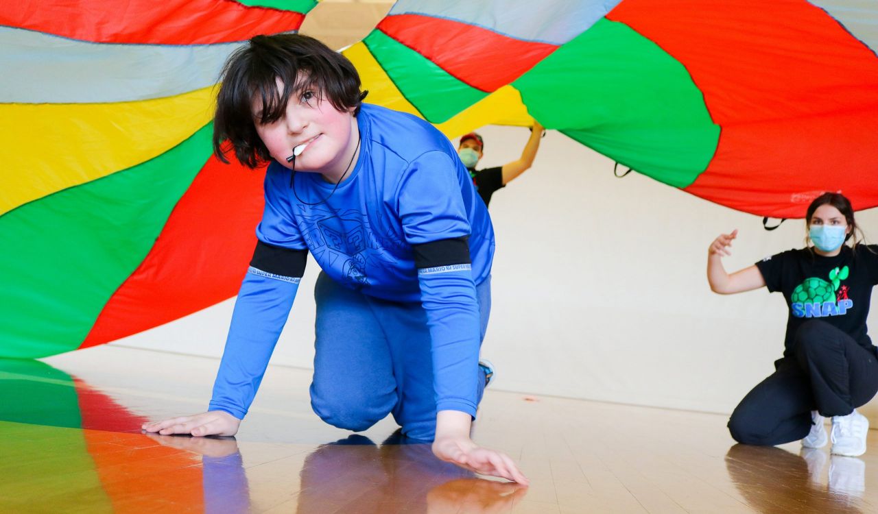 A child crawls on the ground of a gymnasium floor underneath a colourful parachute being used to promote physical activity. Two student volunteers wearing T-shirts with a photo of a turtle and the acronym “SNAP” are in the background. One is holding the parachute, and the other is observing the child.