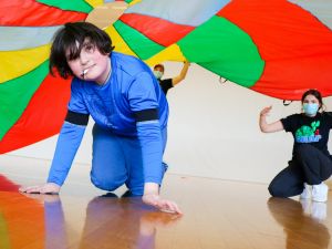 A child crawls on the ground of a gymnasium floor underneath a colourful parachute being used to promote physical activity. Two student volunteers wearing T-shirts with a photo of a turtle and the acronym “SNAP” are in the background. One is holding the parachute, and the other is observing the child.