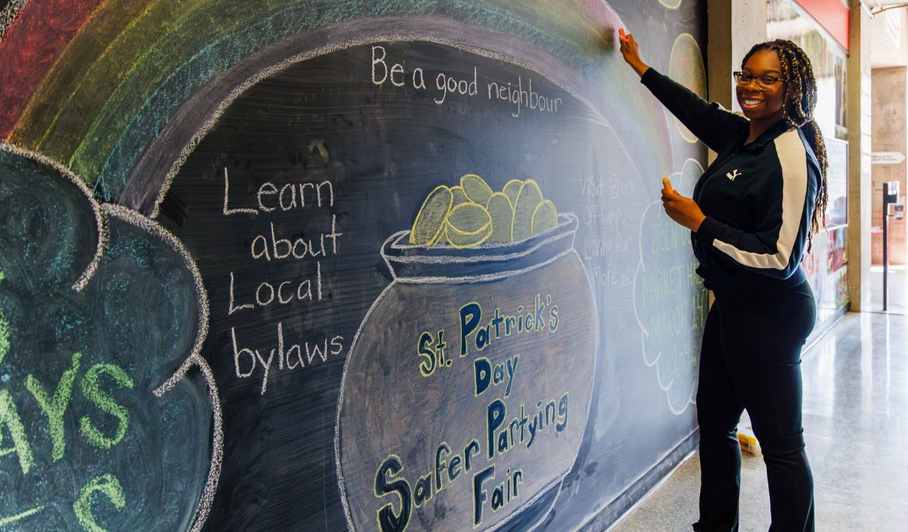 A woman draws a large rainbow over a pot of gold on a chalkboard.