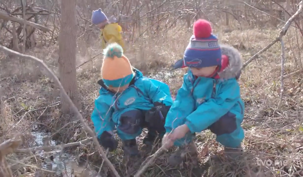 Two small children play with sticks in a forest while wearing winter gear.
