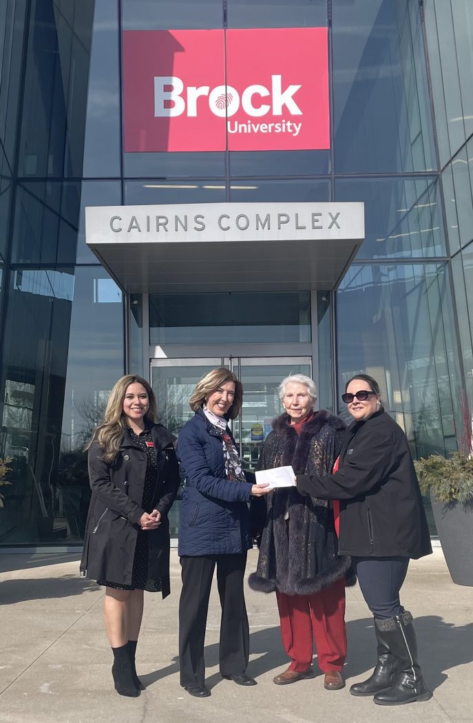 Four women pose with an envelope outside the Cairns Complex building at Brock University on a sunny day.