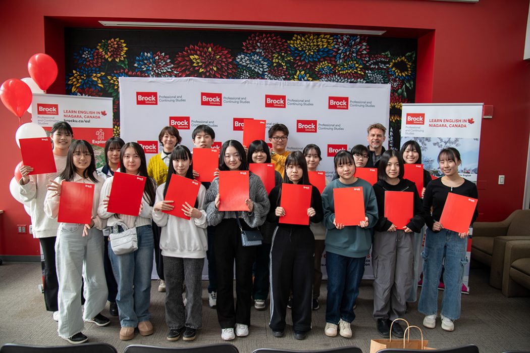 A large group of people standing in front of white backdrop smiling and posing for photo while holding up red folders.