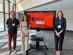 Three university students pose for a photo beside a TV showing a presentation slide.