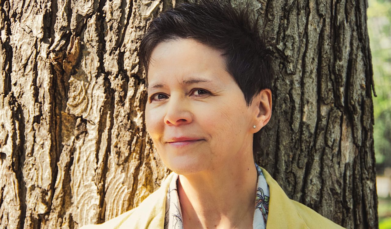 Author Ann-Marie MacDonald shown from the shoulders up, leaning against a tree on a sunny day. She has short, dark hair and wears a pink collared jacket. She is smiling, looking off to the side.