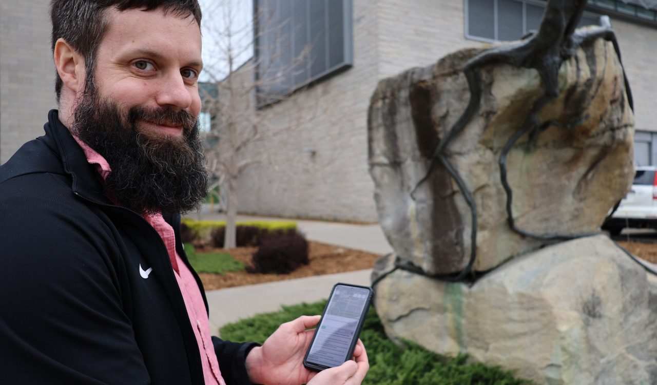 Quinn MacDonald holds a smartphone next to Reinhard Reitzenstein’s sculpture 2000 Years on the Edge on Brock’s main campus.