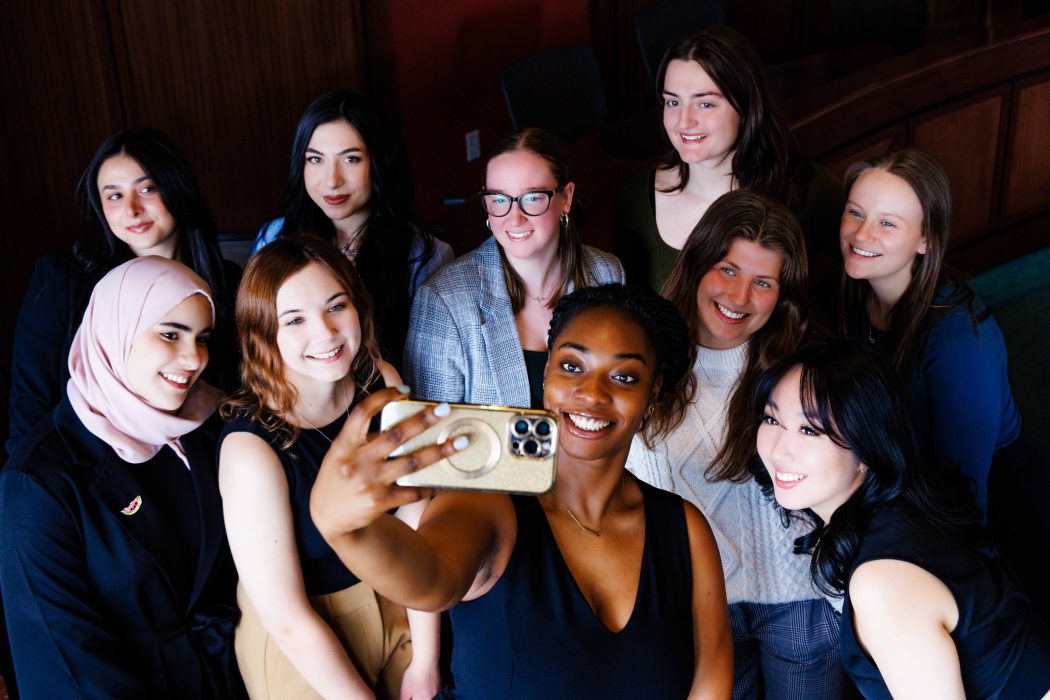A group of 10 women take a selfie with a mobile phone while being illuminated by window light.