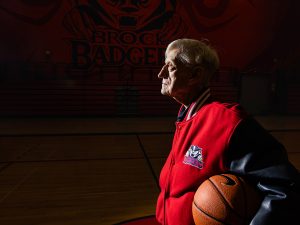 A man stands holding a basketball in a darkly lit gymnasium at Brock University.
