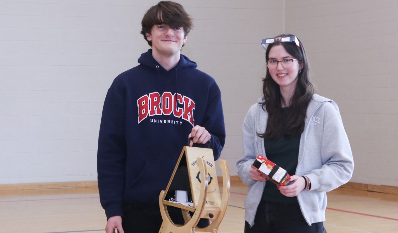 Two Brock University students stand in a gymnasium, one holding a Sun Spotter and the other holding a Ritz cracker box that has been used to create a pinhole camera.