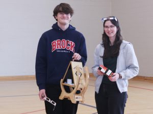Two Brock University students stand in a gymnasium, one holding a Sun Spotter and the other holding a Ritz cracker box that has been used to create a pinhole camera.