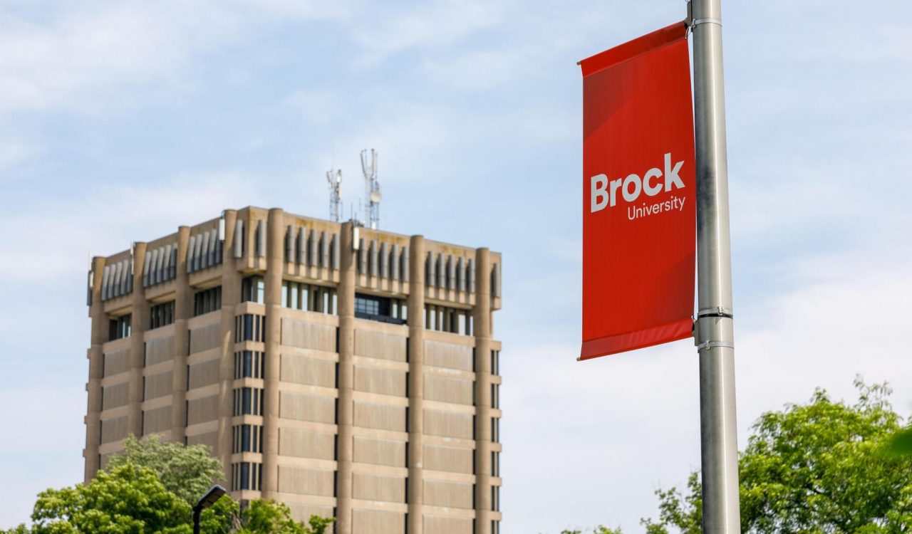 A red Brock University banner hangs from a pole with the Schmon Tower in the background.