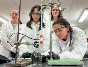 A lab supervisor in a white lab coat observes several students using scientific equipment to add one solution to another drop by drop. The three teenagers are also wearing lab coats.