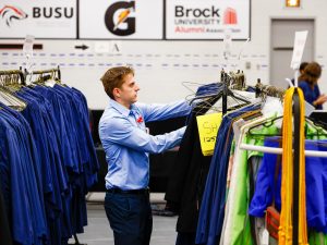 A man stands between two rows of graduation gowns hanging on racks. He is organizing them.