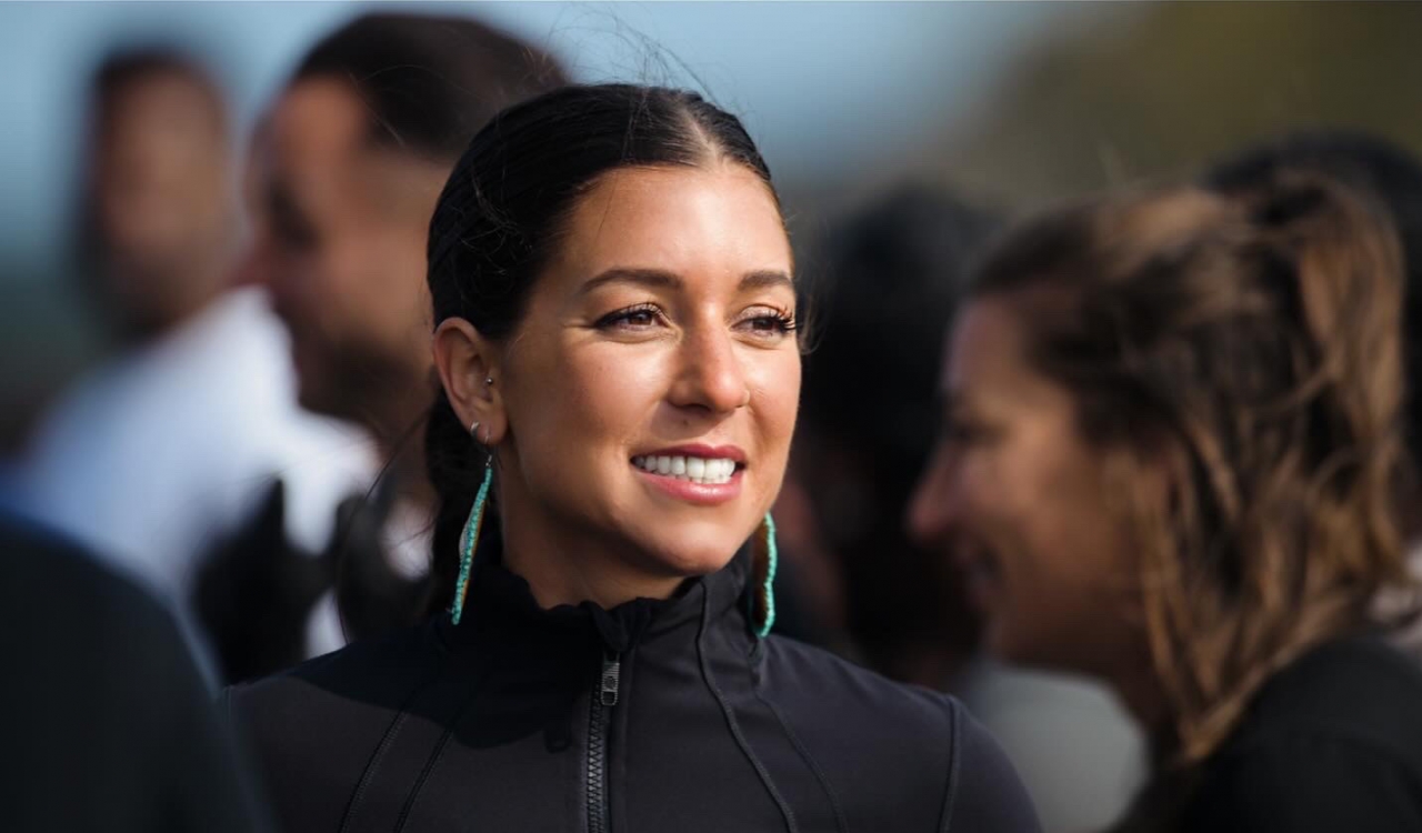 A close up shot of an Indigenous woman shows her dressed in black with turquoise, long earrings. She smiles warmly looking into the distance and the background is blurred behind her. 