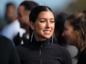A close up shot of an Indigenous woman shows her dressed in black with turquoise, long earrings. She smiles warmly looking into the distance and the background is blurred behind her.