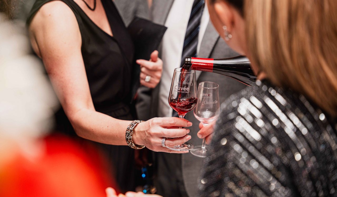 Image of a woman’s hands holding a wine glass that is filled. She is surrounded by a group of people.