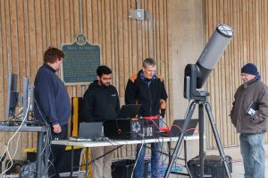 Four people stand around technical equipment used to livestream an event, including laptop computers and a telescope.