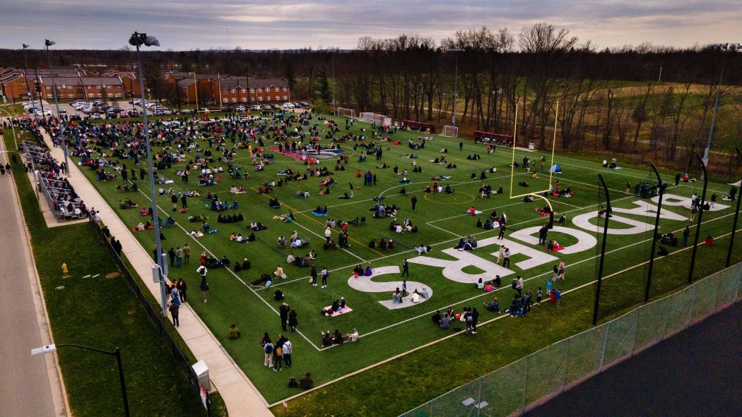 A green turf field with the work BADGERS written in large letters is covered with people sitting and standing.