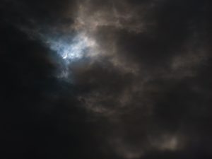 A crescent of the total solar eclipse is seen in the darkened, cloudy sky.