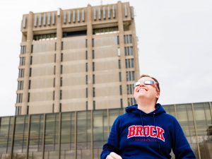 A person wearing a blue Brock hoodie and eclipse glasses looks up toward the sky, with Brock's Schmon Tower visible in the background.