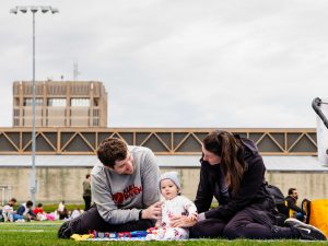 Two adults and a baby sit on a picnic blanket on a football field.