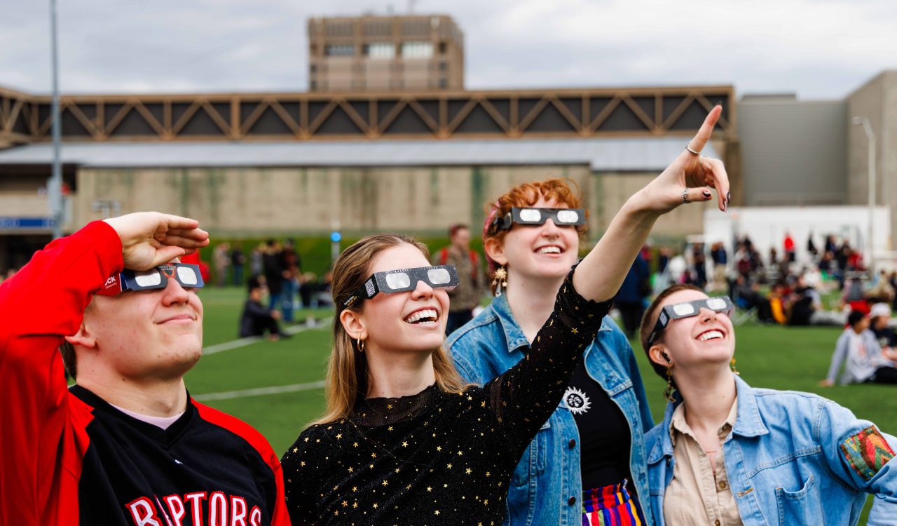 People wearing eclipse glasses stand together on a football field looking up towards the sky. 