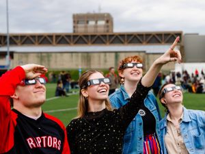 People wearing eclipse glasses stand together on a football field looking up towards the sky.