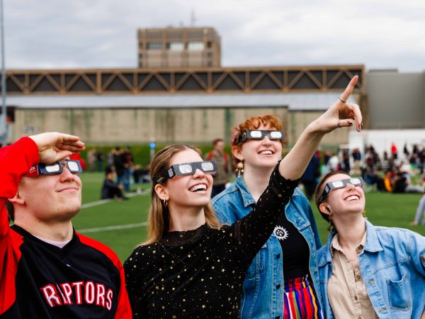 People wearing eclipse glasses stand together on a football field looking up towards the sky.