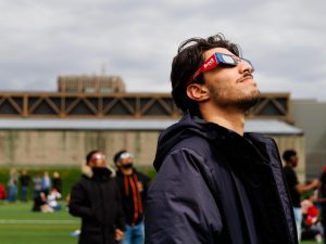 A man wearing Brock-branded eclipse glasses stares up towards the sky.
