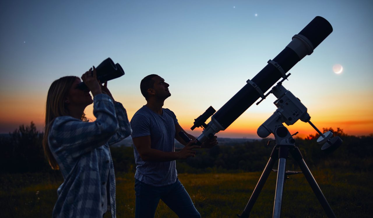 A couple stargazes with a telescope at sunset.