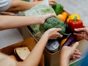 Close-up of two sets of hands placing broccoli and peppers into a cardboard box filled with cans and other food supplies.