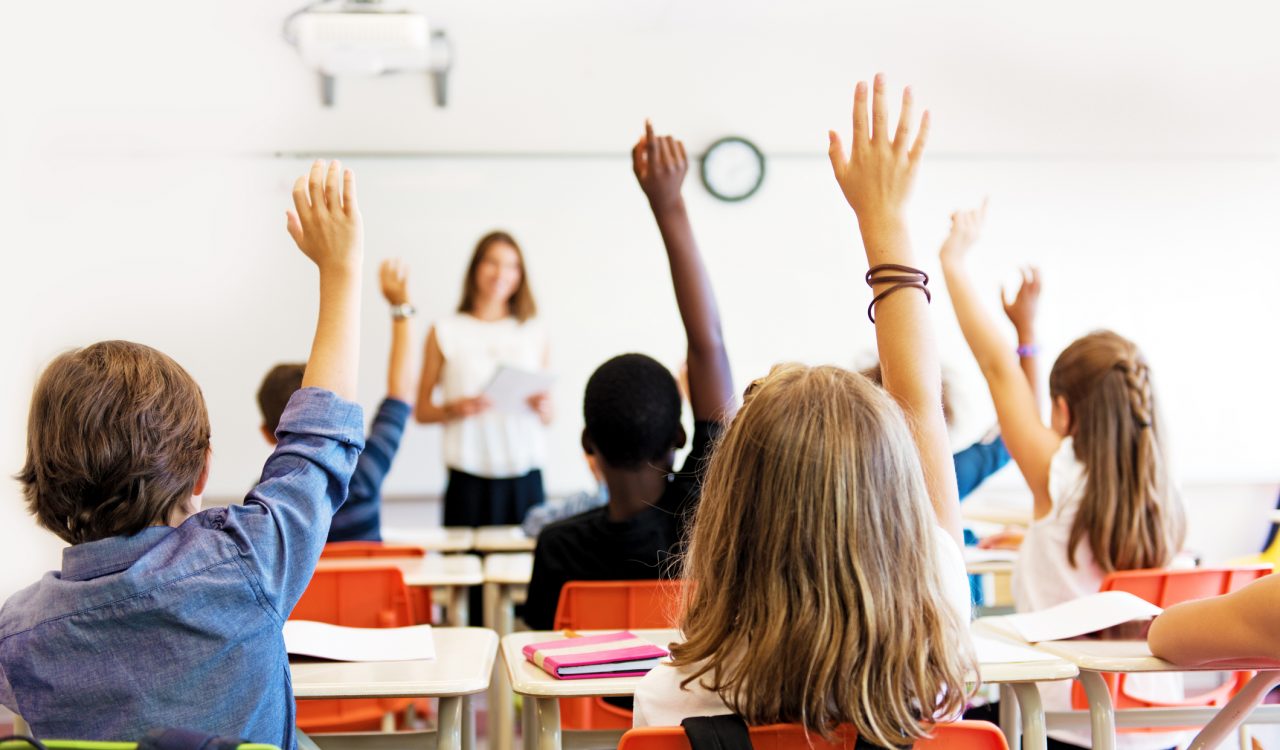 A bright, sunny classroom shows students from behind, holding up their hands while looking at the teacher standing at the front of the classroom.