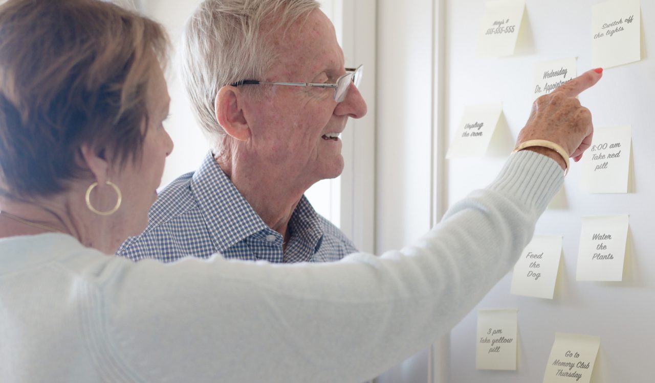 An elderly couple looks at sticky note reminders on a wall.