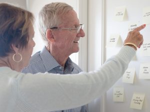 An elderly couple looks at sticky note reminders on a wall.
