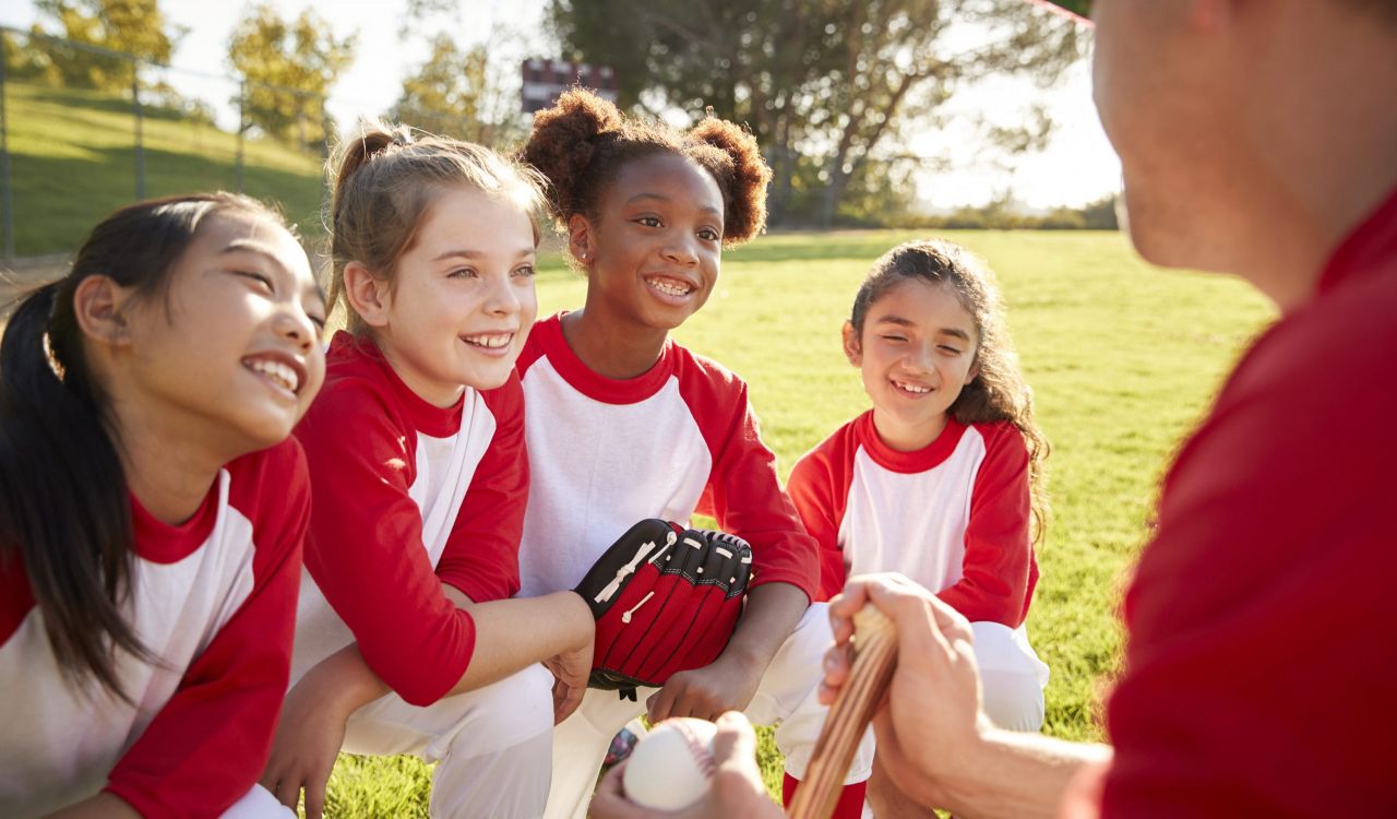 Four girls in red and white baseball uniforms kneel on the grass as their coach speaks with them. One girl is holding a baseball glove. Their coach is holding a baseball. 