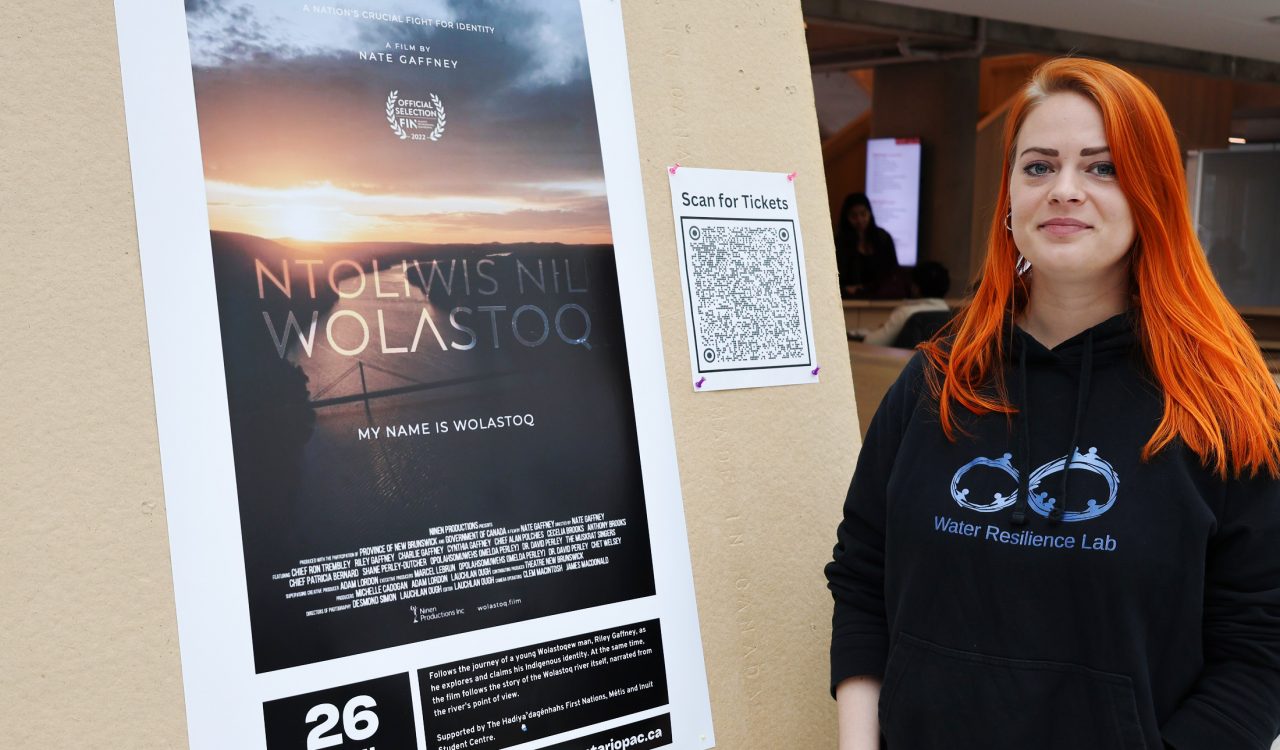 A woman stands in front of a movie poster for the film My Name is Wolastoq.