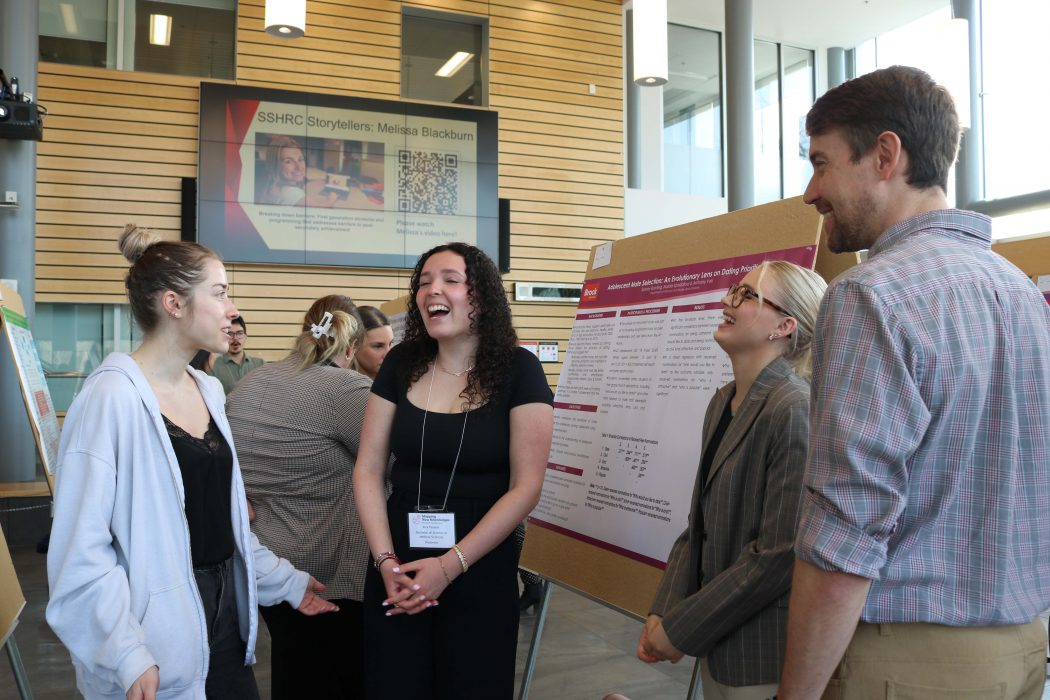 Three university students talk to a professor in front of a research poster at a conference. 