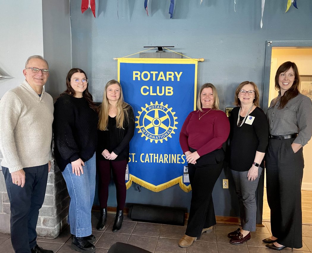 Six people pose side-by-side for a photo next to a Rotary Club banner.