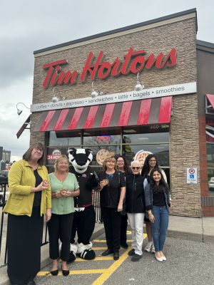 Group of people, and Brock mascot, Boomer, stand in front of a Tim Hortons cafe while smiling and holding Smile Cookies.