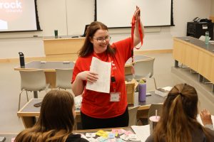 A woman in a red shirt holds a paper in one hand and a lanyard in another as she makes a presentation to room of high school students.