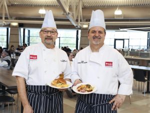 Two chefs in white uniforms stand next to each other in a cafeteria.
