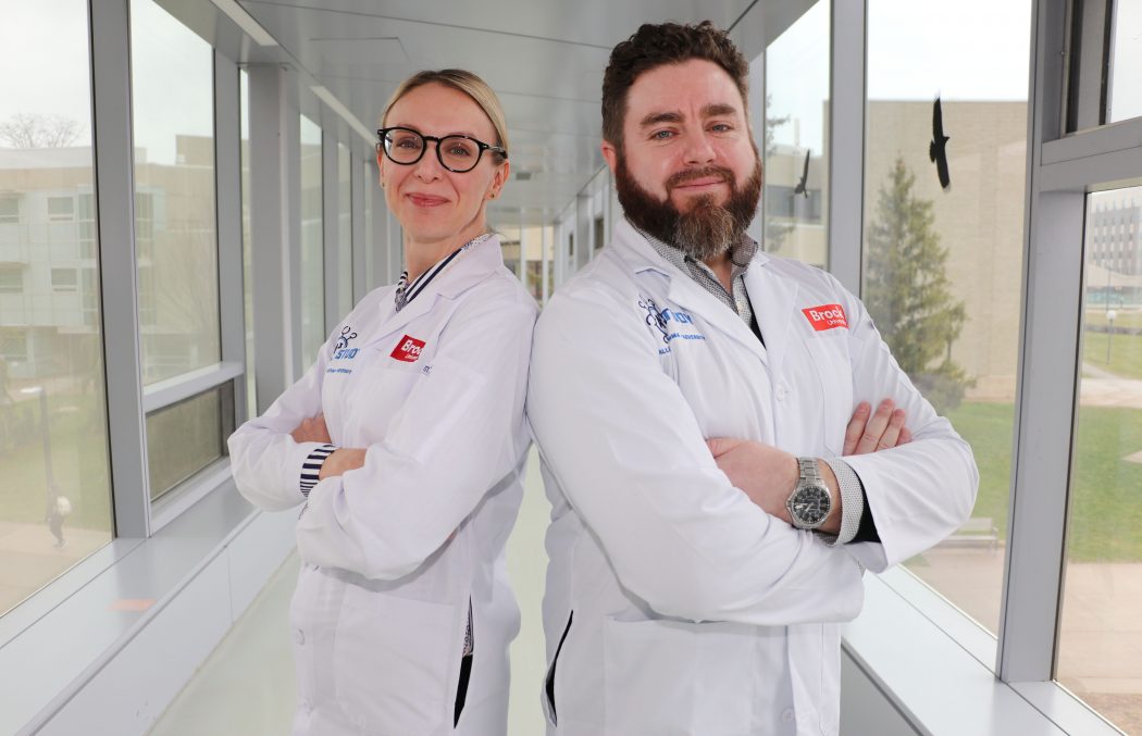 Associate Professors of Health Sciences Karen Patte (left) and Adam MacNeil (right) stand back-to-back, arms crossed and faces smiling and facing the camera, with a blurred hallway bridge in the background.