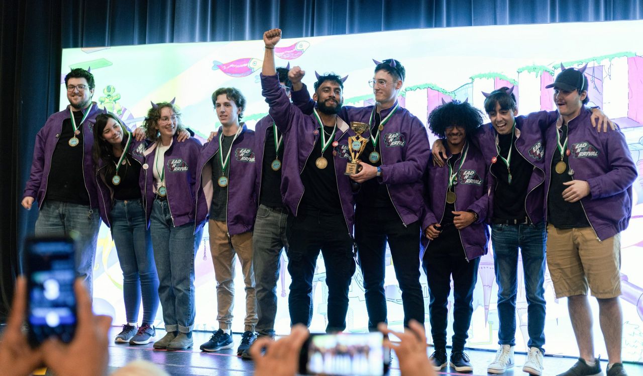 A group of people stand, arms linked, on a stage in front of a white digital screen. The people are wearing matching purple jackets and smiling, each with a medal around their neck. A person in the middle of the group holds a trophy with his arm raised. 