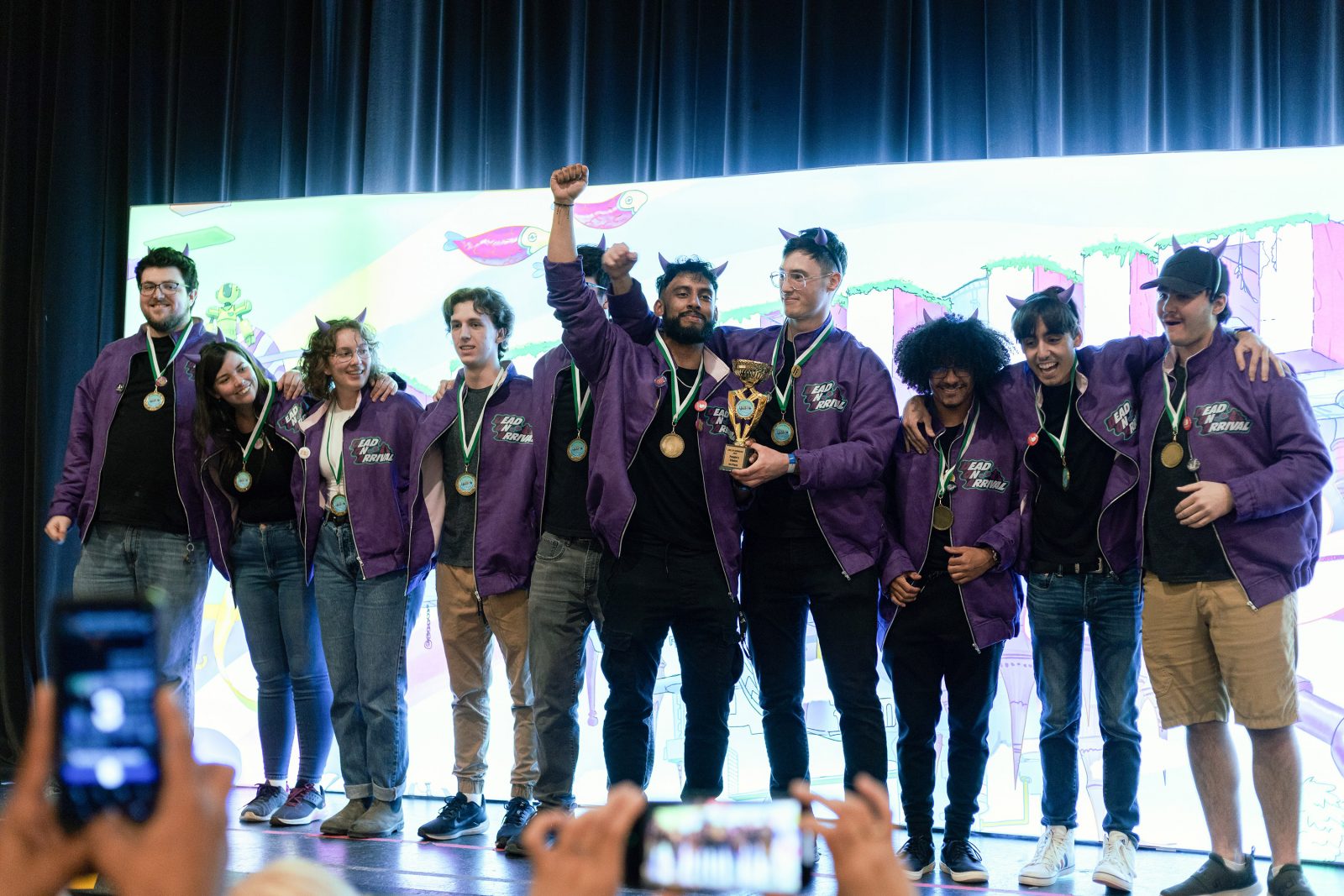 A group of people stand, arms linked, on a stage in front of a white digital screen. The people are wearing matching purple jackets and smiling, each with a medal around their neck. A person in the middle of the group holds a trophy with his arm raised.