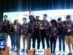 A group of people stand, arms linked, on a stage in front of a white digital screen. The people are wearing matching purple jackets and smiling, each with a medal around their neck. A person in the middle of the group holds a trophy with his arm raised.